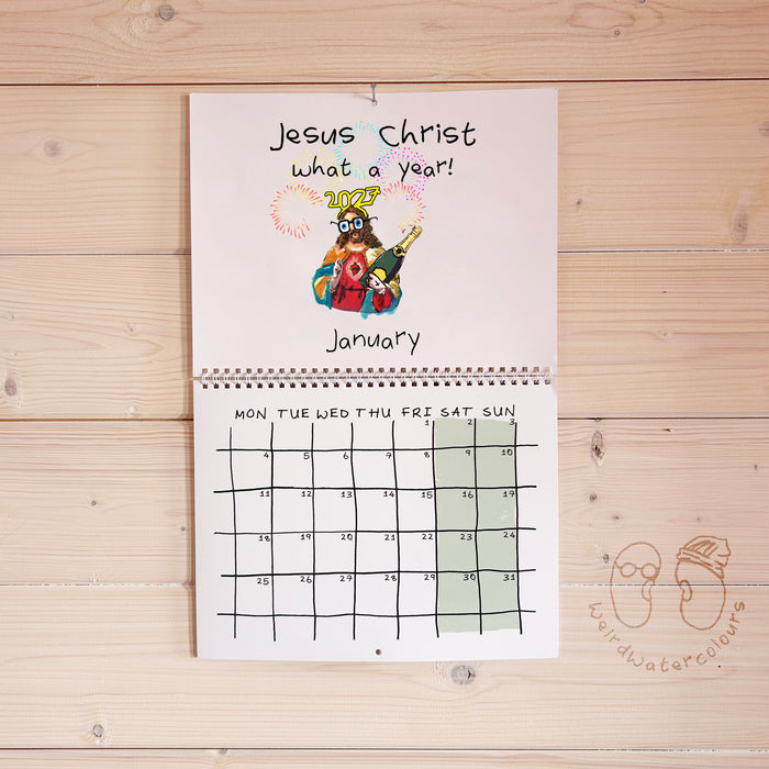 A spiral-bound monthly calendar page titled 'Jesus Christ what a year!' hangs on a light wood plank wall. The top section has a colorful cartoon illustration of Jesus and the word 'JESUS' in gold, with 'January' written below.