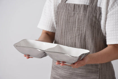 Person holding two white square plates with a gray apron on a light gray background