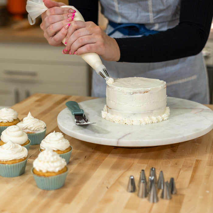 Woman icing a white cake on a white platter on a wooden counter next to vanilla cupcakes