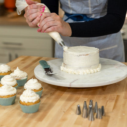 Woman icing a white cake on a white platter on a wooden counter next to vanilla cupcakes