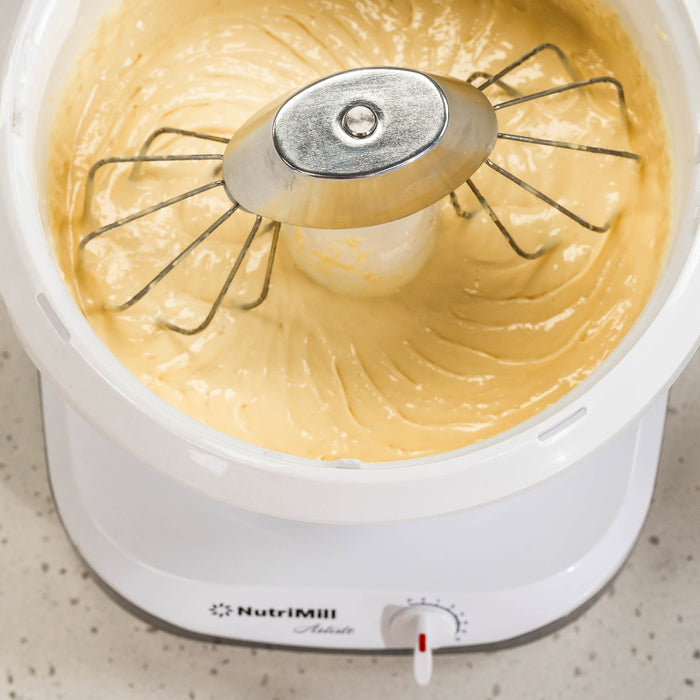 A NutriMill artiste mixer mixing a yellow batter with wire whips and a metal whip driver