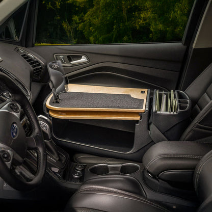 A car interior featuring the AutoExec GripMaster Car Desk with Mega Grip™ Drill-Base Mount attached to the passenger seat, complete with a desk mat and document organizer next to the seat, set against a backdrop of green trees seen through the window.
