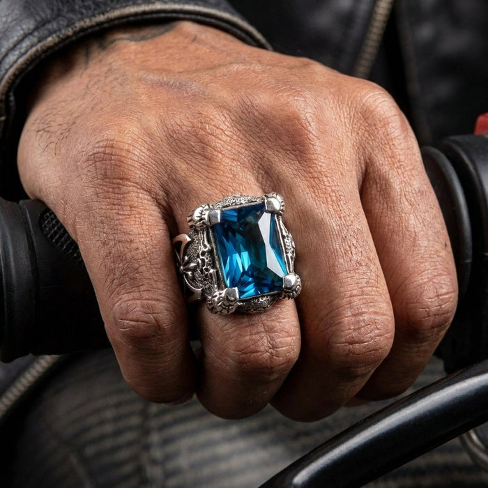 Close-up of a man's hand gripping a motorcycle handlebar while wearing a chunky sterling silver dragon claw ring featuring a large rectangular blue topaz gemstone.