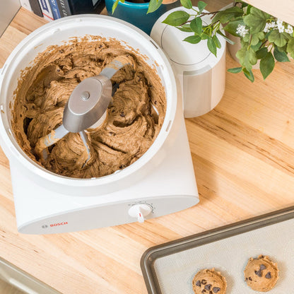 Bosch white universal plus mixer mixing cookie dough with cookie paddles on light wooden surface next to a tray of cookie dough