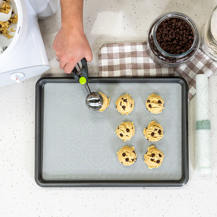 Person scooping chocolate chip cookie dough on cookie sheet next to a jar of chocolate chips on a white speckled counter