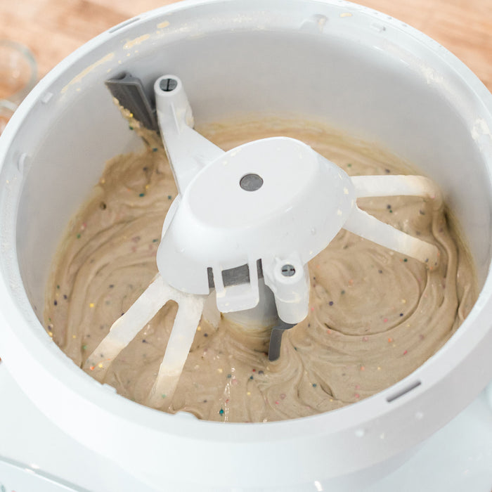 A white Bosch universal plus mixer mixing white cake batter with the NutriMill cake paddles and bowl scraper