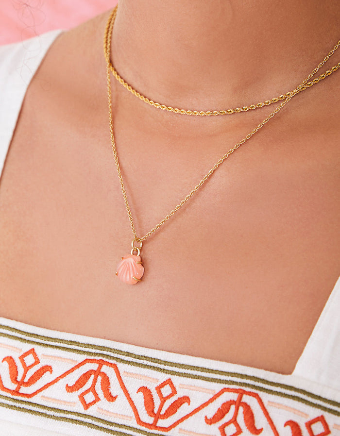 Close-up of a person wearing a gold necklace with a shell pendant, against a blurred background.