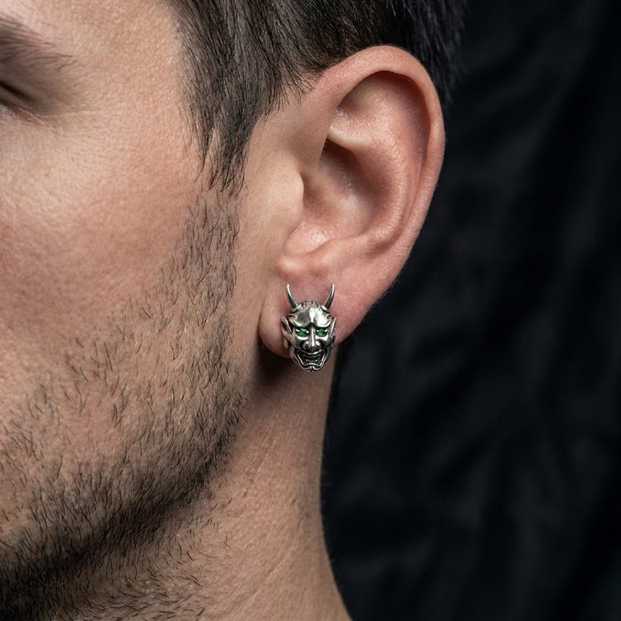 Close-up profile of a man wearing a silver devil skull stud earring featuring green gemstone eyes, set against a dark background.