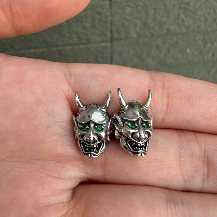 A pair of sterling silver Oni mask earrings with piercing green CZ eyes held in a hand.