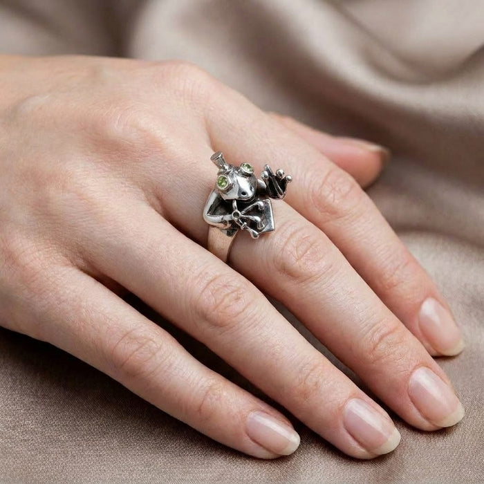 Close-up of a hand wearing a detailed sterling silver frog ring featuring a tiny top hat and bright green gemstone eyes, resting against a soft beige silk background.