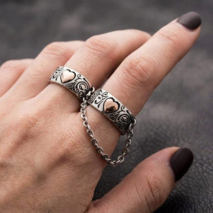 Close-up of a hand with dark nail polish wearing a gothic silver two-finger ring connected by a chain, featuring ornate filigree engravings and rose gold heart accents, set against a dark textured background.