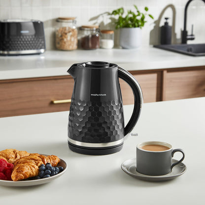 Black electric kettle on a kitchen counter with a plate of food and a cup of coffee.