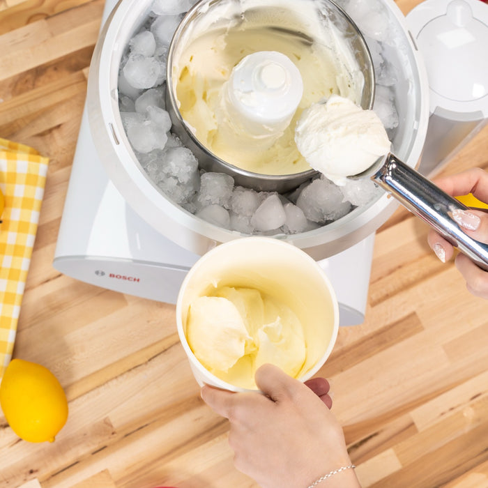 Vanilla ice cream being scooped from a ice cream maker attachment into a white container next to a lemon on a wooden surface.