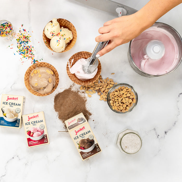 A woman scooping strawberry ice cream into a waffle cone next to more ice cream, ice cream mix, and toppings on a white counter