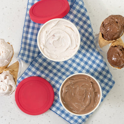 A container filled with strawberry ice cream and a container filled with chocolate ice cream on a blue checkered cloth on a white counter next to chocolate and strawberry ice cream in cones
