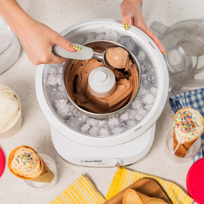 Lifestyle photo of hands using a white ice cream maker with chocolate ice cream, cones, and dishes nearby