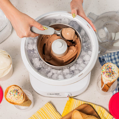 Lifestyle photo of hands using a white ice cream maker with chocolate ice cream, cones, and dishes nearby