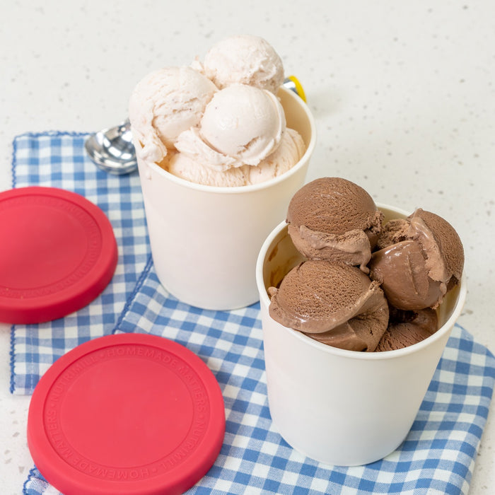 Two white containers with red lids filled with vanilla and chocolate ice cream on a blue checkered tablecloth on a white counter