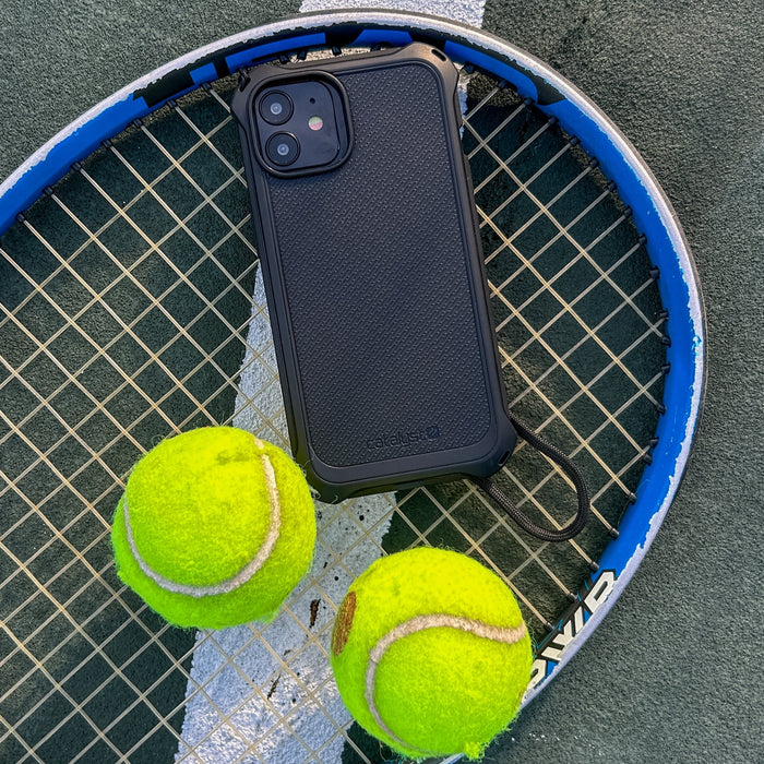 iPhone protected by a Catalyst Drop Proof Textured Case in black is positioned on a tennis racket with two tennis balls close by on the court.