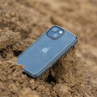 A Catalyst Case US iPhone 14 Plus - Influence Case, black with visible Apple logo, lies face down in loose brown dirt, partially covered by soil. The camera lenses and flash remain clearly visible on the raised top corner.