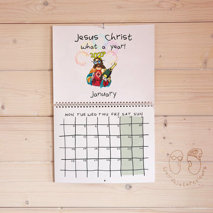 A spiral-bound monthly calendar page titled 'Jesus Christ what a year!' hangs on a light wood plank wall. The top section has a colorful cartoon illustration of Jesus and the word 'JESUS' in gold, with 'January' written below.