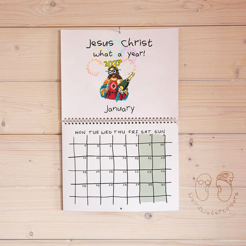 A spiral-bound monthly calendar page titled 'Jesus Christ what a year!' hangs on a light wood plank wall. The top section has a colorful cartoon illustration of Jesus and the word 'JESUS' in gold, with 'January' written below.