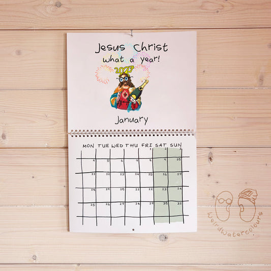 A spiral-bound monthly calendar page titled 'Jesus Christ what a year!' hangs on a light wood plank wall. The top section has a colorful cartoon illustration of Jesus and the word 'JESUS' in gold, with 'January' written below.