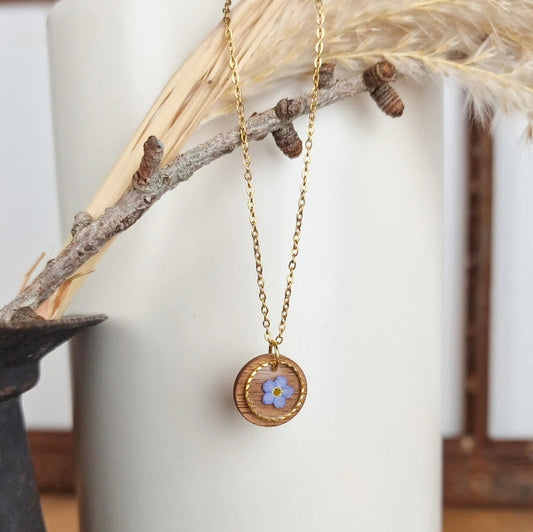 Goldfarbene Edelstahlkette mit rundem Anhänger aus Nussholz, eingefasst mit feinem Metallring und echter blauer Vergissmeinnicht-Blüte im Resin, dekorativ an einem Zweig arrangiert.