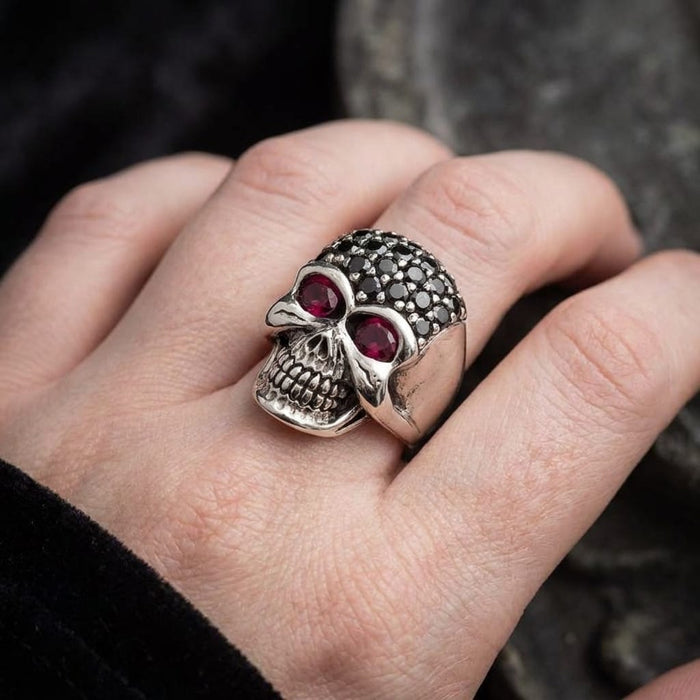 Close-up of a person's hand wearing a gothic silver lady skull ring featuring black crystal pave on the top of the skull and striking red stones in the eye sockets, set against a dark background.