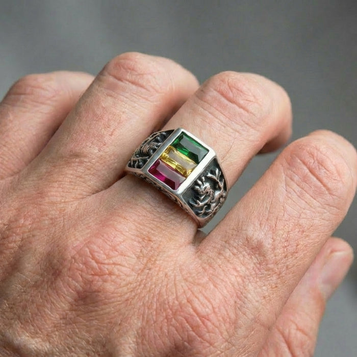 Close-up of a sterling silver Lion of Judah ring worn on a man's finger, featuring a rectangular face with vertical green, yellow, and red gemstones and intricate lion relief carvings on the band.