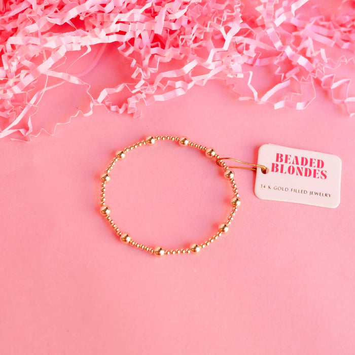 A gold-colored beaded bracelet displayed on a pink background with crinkled pink paper to the sides and a tag attached to the bracelet.