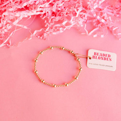 A gold-colored beaded bracelet displayed on a pink background with crinkled pink paper to the sides and a tag attached to the bracelet.