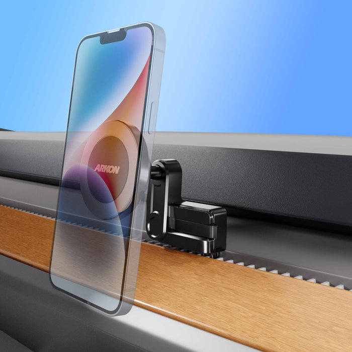 A transparent smartphone is mounted on a black and silver clamp attached to the edge of a wood and metal surface, set against a blue gradient background—an ideal setup for showcasing the Magnetic Phone Vent Mount for Tesla Model 3 (2017–2022) & Model Y (2020–2023), a must-have accessory that features a secure magnetic vent mount.