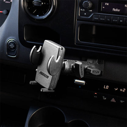 A black Mega Grip™ Phone Drill Base Mount with a double socket swivel arm is attached to a car’s dashboard vent, positioned below the radio controls and air conditioning dials, in a dimly lit car interior.