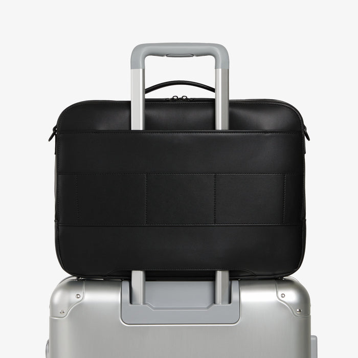 Carbon Black (Vegan Leather) | Back view of Metro Briefcase in Carbon Black with trolley sleeve attached to a silver suitcase handle