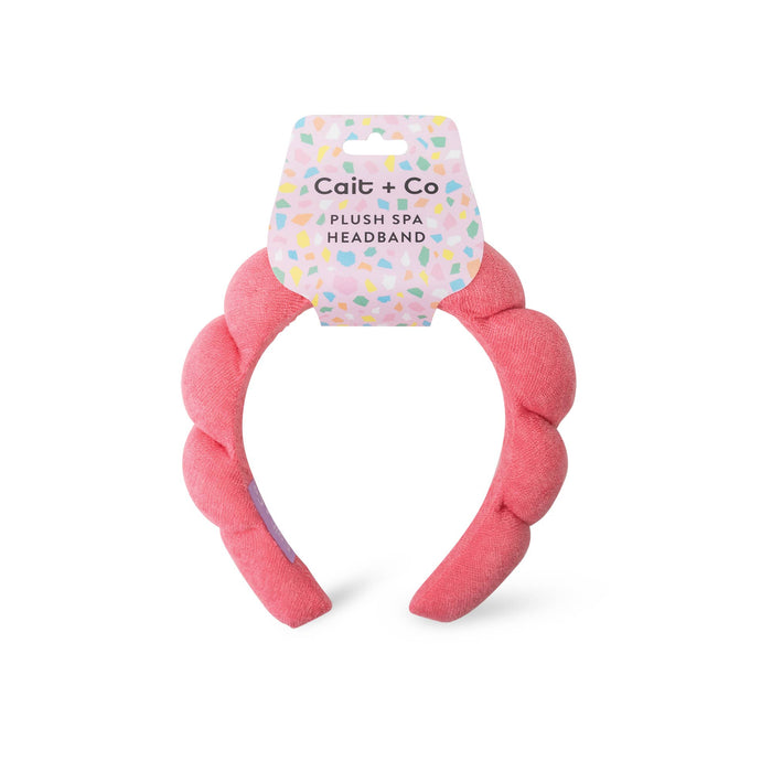 A pink terry cloth headband with a brand tag that reads 'cait + co. plush spa headband'.