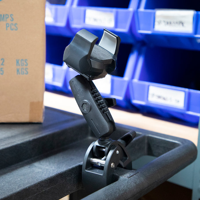 The RoadVise® Ultra Holder with Clamp Mount, in black, is attached to a carts edge. Blue plastic storage bins and a cardboard box are visible in the background.
