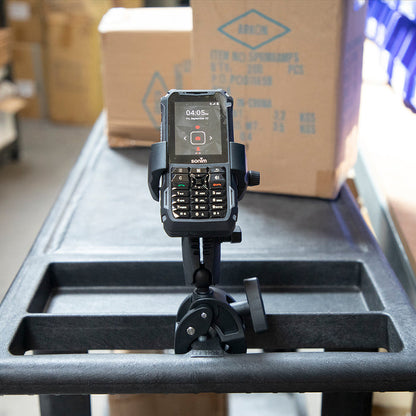 A black handheld device with a physical keypad is mounted in the RoadVise® Ultra Holder with Clamp Mount on a rolling cart in a warehouse, surrounded by boxes and shelves.