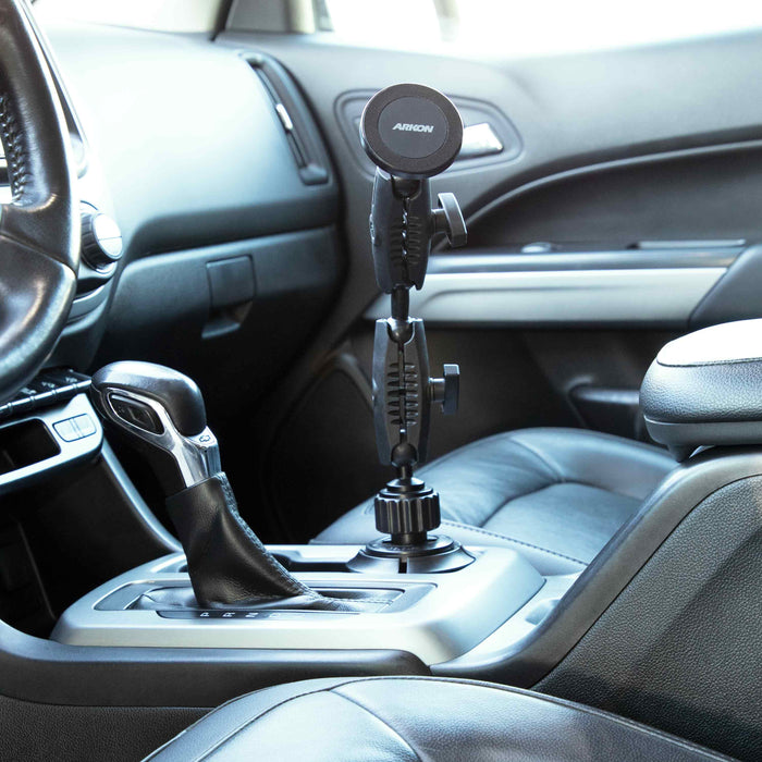 An Arkon Mounts Robust® Dual Shaft Cup Holder Mount with Magnetic Phone Holder (25mm, MagSafe compatible) is installed in the car’s center console cup holder next to the gear shift, with black leather seats and a modern dashboard in view.