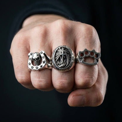 A man's hands wearing a collection of heavy gothic rings, featuring the large Roman numeral signet ring.