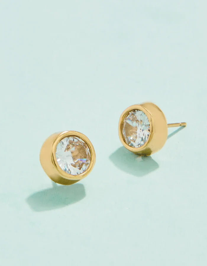 A pair of gold-colored stud earrings with cubic zirconia crystals on a light background.