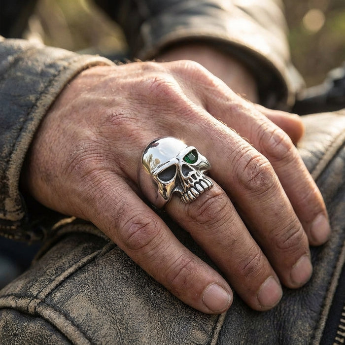 Close-up of a masculine hand wearing a polished sterling silver skull ring featuring a single green gemstone eye, resting on a distressed brown leather biker jacket.