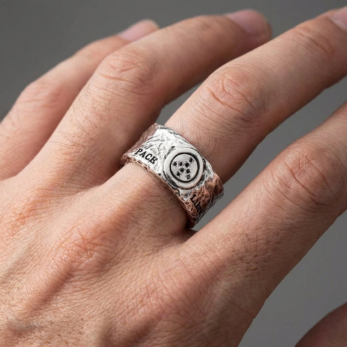Close-up of a man's hand wearing a chunky, distressed silver peace ring featuring a circular emblem and stamped lettering on a rugged, textured band, set against a soft neutral background.
