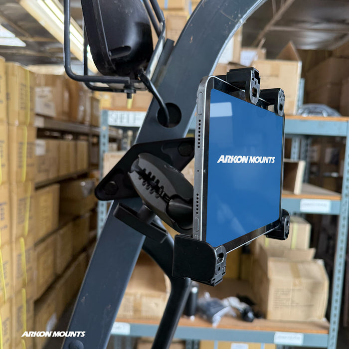 A tablet is mounted on a forklift using the XMS™ Heavy Duty Forklift Mount with LockVise™ Locking Tablet Holder, as part of a reliable warehouse tablet mounting system, in a warehouse filled with cardboard boxes on metal shelves.