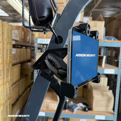 A tablet is mounted on a forklift using the XMS™ Heavy Duty Forklift Mount with LockVise™ Locking Tablet Holder, as part of a reliable warehouse tablet mounting system, in a warehouse filled with cardboard boxes on metal shelves.