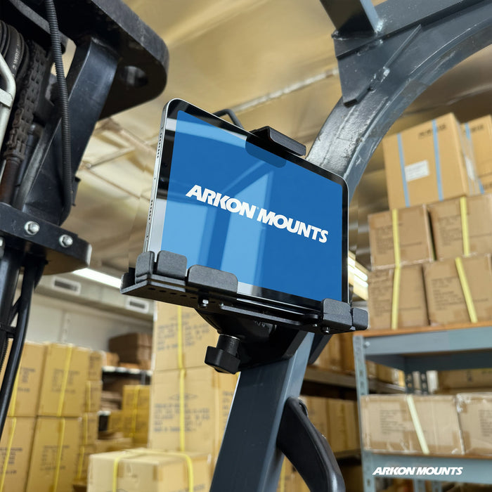 A tablet is securely held in the XMS™ Heavy Duty Forklift Mount with LockVise™ Metal Locking Tablet Holder, which is attached to warehouse equipment and surrounded by shelves and stacks of cardboard boxes.
