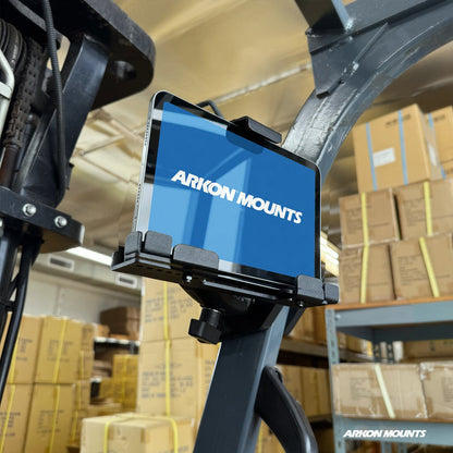 A tablet is securely held in the XMS™ Heavy Duty Forklift Mount with LockVise™ Metal Locking Tablet Holder, which is attached to warehouse equipment and surrounded by shelves and stacks of cardboard boxes.